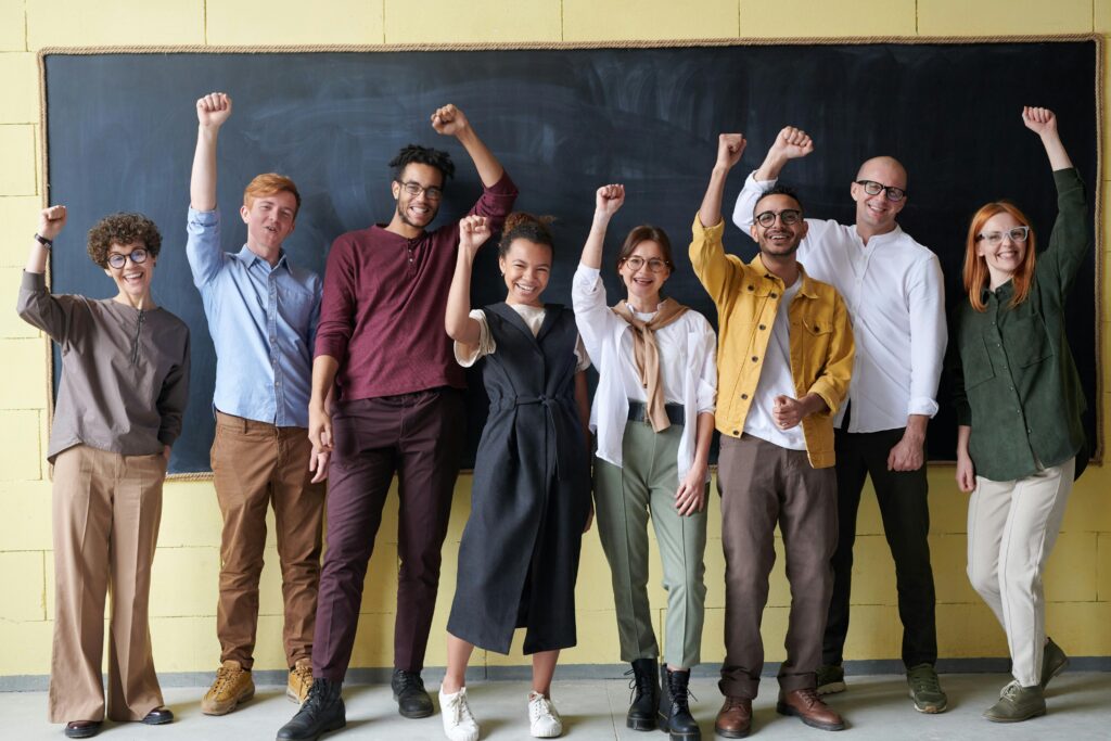 pexels-photo-3184432-3184432 A group of eight adults cheerfully standing with raised fists in front of a blackboard indoors.